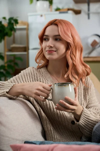 Parlak saçlı kadın rahat bir kanepede dinlenirken sıcak bir içecek yudumluyor, yavaşça gülümsüyor..