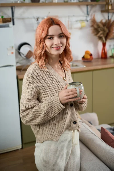 Sıcak güneş ışığı mutfağı dolduruyor. Genç bir kadın içkisini tutuyor ve nazikçe gülümsüyor..