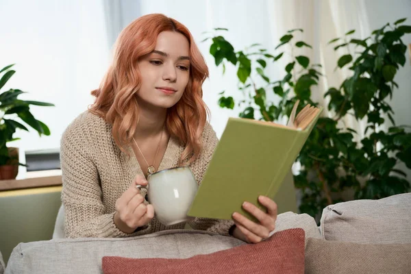 Şık saçlı genç bir kadın rahat bir koltukta dinleniyor, kitabına dalmış çay yudumlarken..