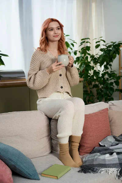 Genç bir kadın, bitkilerle çevrili kanepede rahatça dinlenirken çaydan zevk alır..