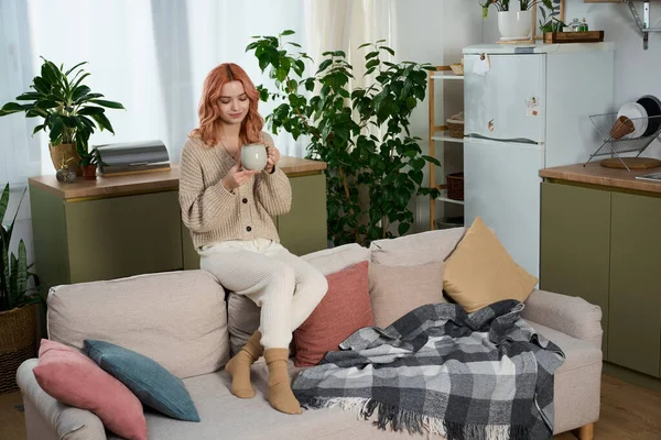 Güzel genç bir kadın şık yaşam alanında dinlenirken rahatlatıcı bir fincan çayın tadını çıkarıyor..