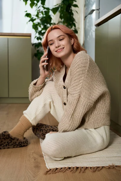 Genç bir kadın, rahat bir atmosferde telefonda konuşurken gülümseyerek yerde oturuyor..