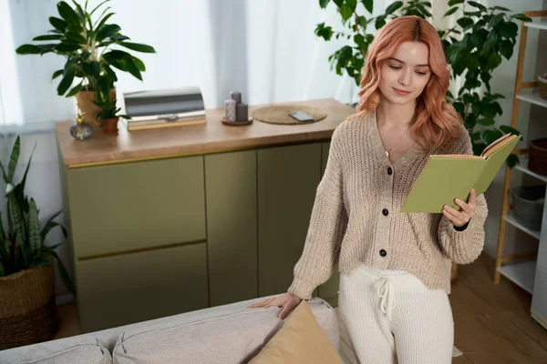 Güzel saçlı genç bir kadın bitkilerle dolu, rahat ve şık bir odada sessizce okuyor..