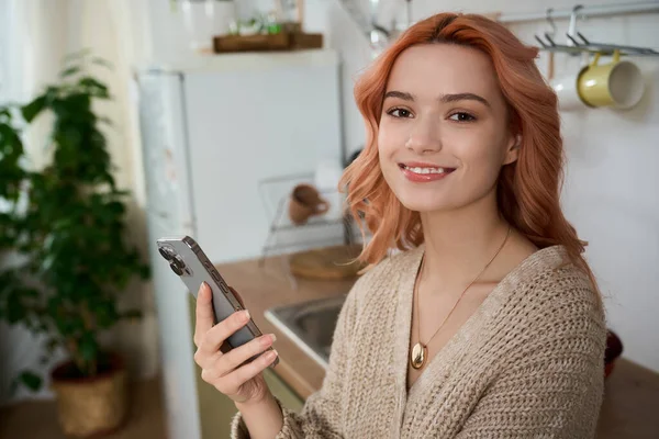 Güzel genç bir kadın, akıllı telefonunu sıcak bir mutfak ortamında tutarken hoş bir şekilde gülümsüyor..