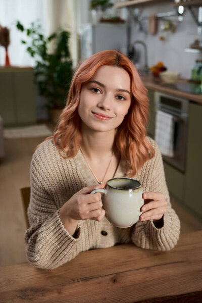 A young woman smiles with a warm cup at a wooden table in her cozy kitchen.