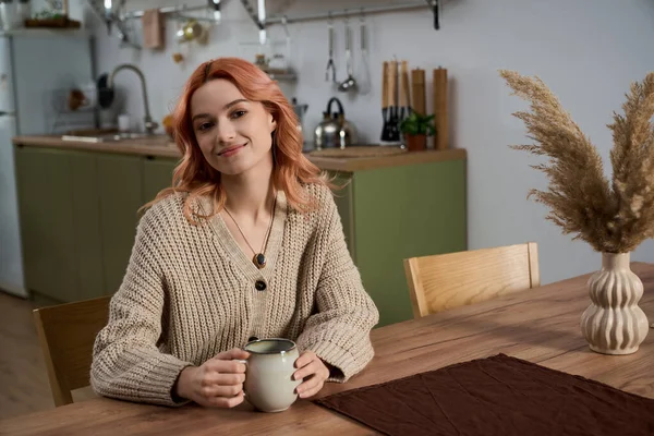 Güzel genç bir kadın ahşap bir masada gevşiyor, şık bir mutfakta sıcak içkisinin tadını çıkarıyor..