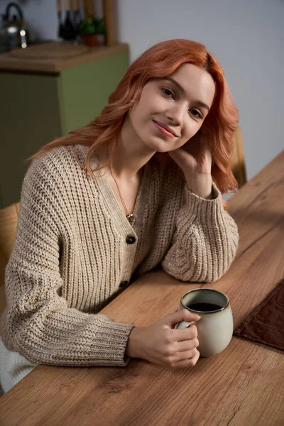 Güzel genç bir kadın ahşap bir masada oturup sıcak bir içeceğin tadını çıkarıyor ve içten bir gülümsemeyle gülümsüyor..