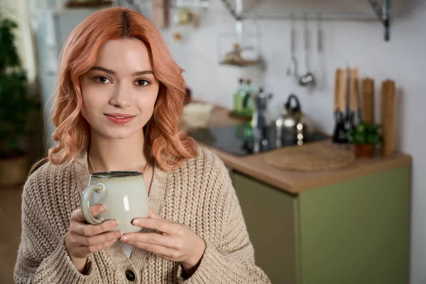 Parlak bir mutfakta, genç ve güzel bir bayan sıcak ve neşe saçan dumanı tüten bir fincan kahve tutar..