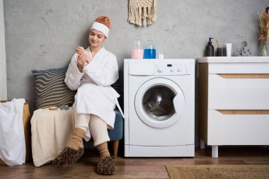 Rahat bir bornoz giymiş genç bir kadın çamaşır odasında oturmuş telefonunu kontrol ediyor..