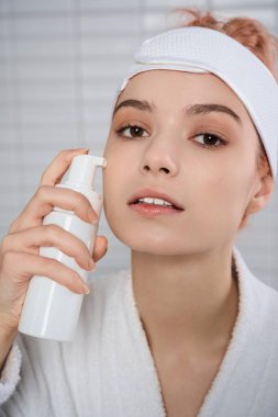 Saç bandı takan genç bir kadın banyodaki parlak cildine nemlendirici sürüyor..