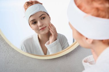 Güzel genç bir kadın, yuvarlak bir aynaya bakarken nazikçe yüzüne dokunur..