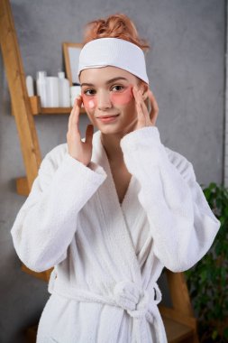 Neşeli genç bir kadın rahatlatıcı bir spa ortamında dinlenirken cilt bakım ürünleri uyguluyor..