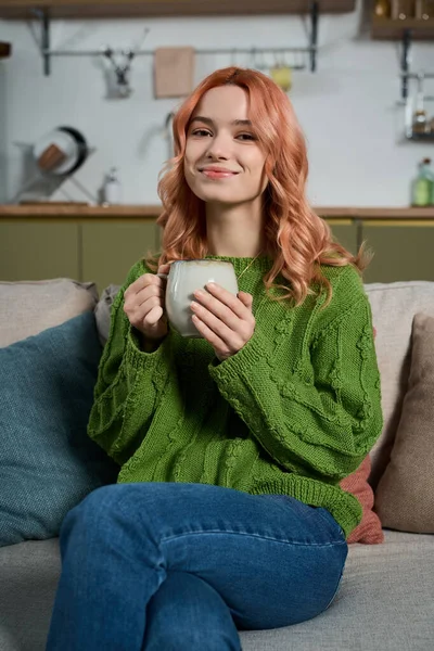 Güzel saçlı genç bir kadın rahat yaşam alanında dinlenirken sıcak bir içeceğin tadını çıkarıyor..