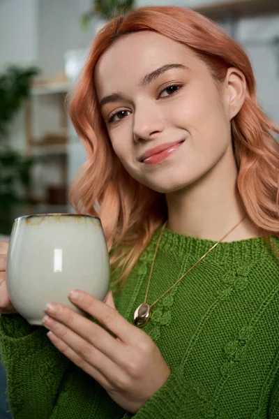 Pembe saçlı genç bir kadın sıcak bir içecek tutarken nazikçe gülümsüyor, neşe saçıyor..