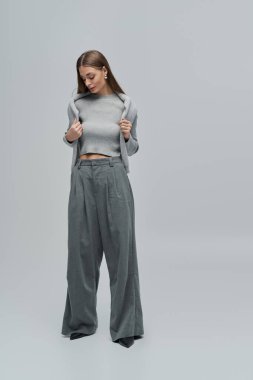 Tarz sahibi genç bir kadın, yumuşak bir arkaplana karşı parlak gri giysiler içinde güven içinde poz verir..