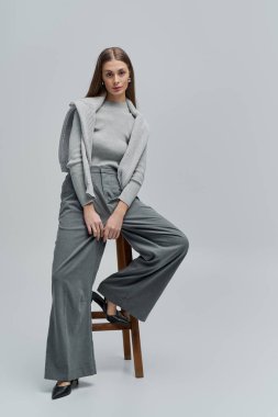 Parlak ışıklandırılmış stüdyoda, kendine güveni tam, şık, gri kıyafetli güzel bir genç kadın sergileniyor..