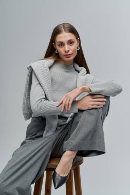 Tabureye oturmuş zarif genç bir kadın zarif bir ifadeyle gri bir elbiseyle zarafet yayıyor..