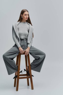 Gri giyinmiş şık genç bir kadın tabureye oturmuş özgüven ve zarafeti gözler önüne seriyor..