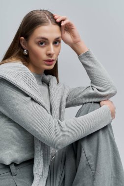 Güzel genç bir kadın zarif bir şekilde oturur, şık kıyafetini minimalist bir ortamda sergiler..