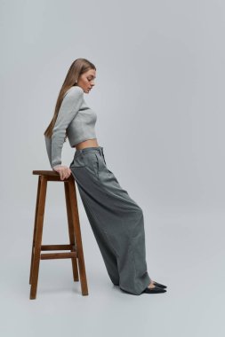 Şık giyinmiş genç bir kadın tabureye yaslanıyor, kendine güven ve cazibe saçıyor..