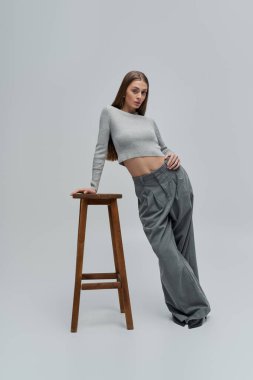 Güzel genç bir kadın zarif bir şekilde ahşap bir taburede duruyor, şık kıyafetini ve duruşunu sergiliyor..