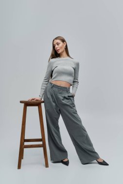 Genç bir kadın, zarif bir şekilde, gri elbisesiyle sade, ahşap bir taburede duruyor..