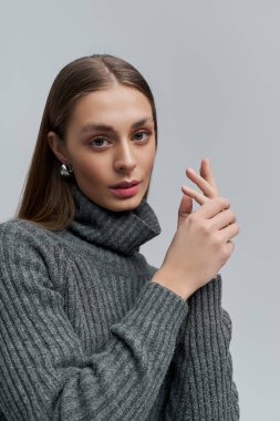 Genç kadın zarif poz veriyor, rahat gri bir balıkçı yaka giyiyor, zarafet ve sıcaklık saçıyor..