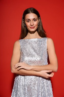 Işıldayan gümüş elbiseli genç bir kadın, koyu kırmızı bir arka plana karşı zarafet sergiliyor..