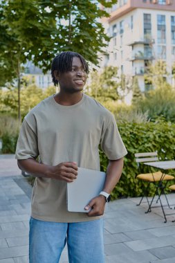 Mutlu bir genç adam güneşli bir öğleden sonra elinde dizüstü bilgisayarla canlı bir parkta yürüyor..