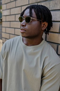 Yakışıklı genç bir adam tuğladan bir duvara yaslanmış şık güneş gözlüğü takıyor ve kendinden emin..