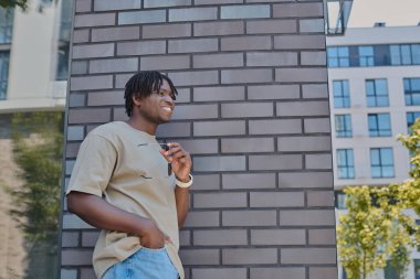 Yakışıklı bir genç adam tuğla bir duvara yaslanırken gelişigüzel gülümsüyor, çevresinin tadını çıkarıyor..