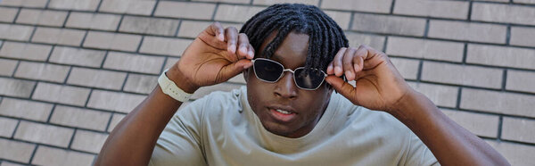 Handsome young man stands outdoors, playfully adjusting his sunglasses with a charming smile.