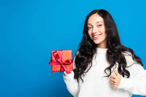Joyful brunette woman showing thumb up and holding christmas present isolated on blue — Stock Photo