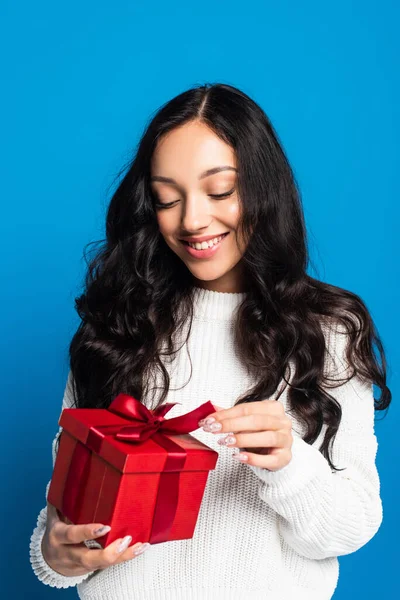 Happy woman in sweater touching ribbon on christmas present isolated on blue — Stock Photo