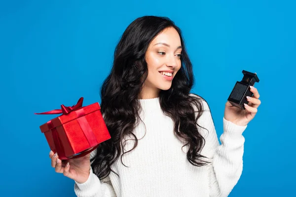 Felice donna in maglione che tiene scatola regalo di Natale e guardando bottiglia con profumo isolato su blu — Foto stock