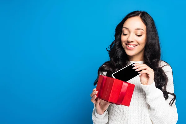 Femme heureuse tenant boîte cadeau de Noël et regardant smartphone avec écran blanc isolé sur bleu — Photo de stock