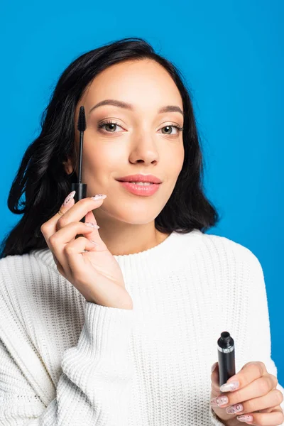 Happy brunette woman holding mascara brush isolated on blue — Stock Photo