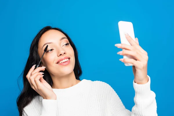 Радостная брюнетка женщина применяя тушь и глядя на смартфон изолирован на синий — стоковое фото