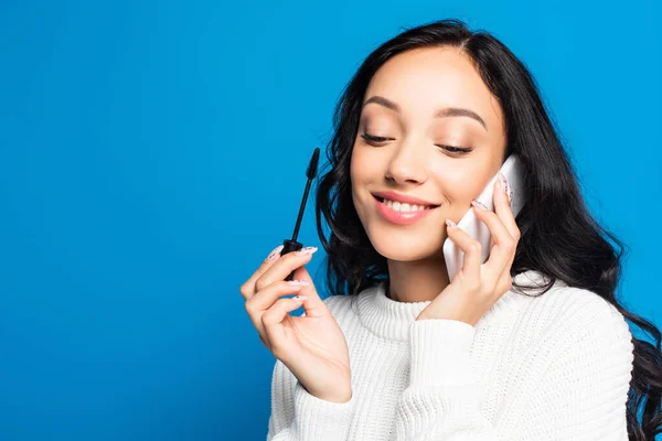 Joyful brunette woman holding mascara brush and talking on smartphone isolated on blue — Stock Photo