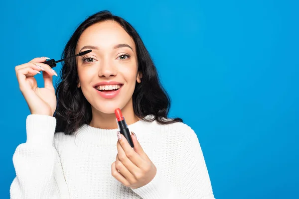 Donna bruna sorridente che applica mascara e tiene il rossetto isolato sul blu — Foto stock