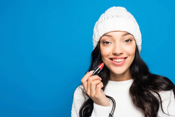 Happy brunette woman in hat holding lipstick isolated on blue — Stock Photo