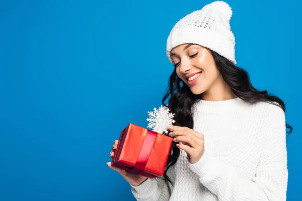 Mujer feliz en sombrero de punto con copo de nieve decorativo cerca de la caja de regalo aislado en azul - foto de stock