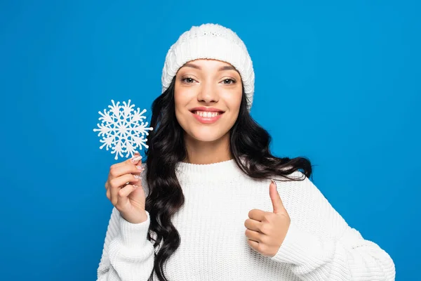 Mulher feliz em chapéu de malha segurando floco de neve decorativo e mostrando polegar para cima isolado em azul — Fotografia de Stock