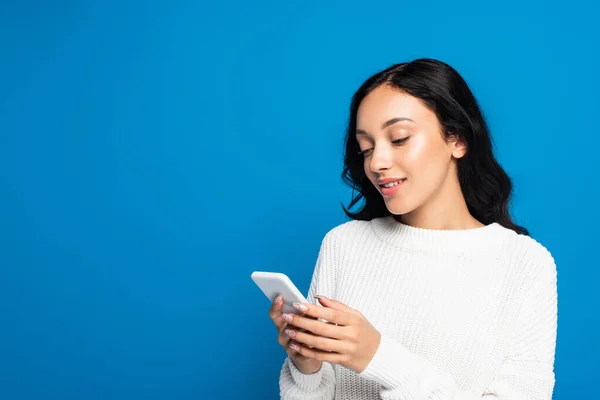 Mujer joven en suéter cálido usando teléfono inteligente aislado en azul - foto de stock