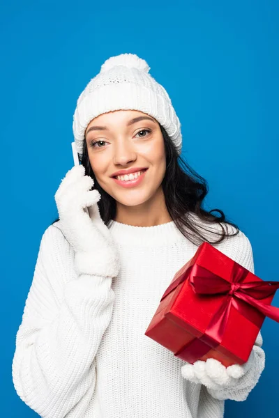 Mujer complacida en sombrero de punto y guantes sosteniendo caja de regalo y hablando en teléfono inteligente aislado en azul - foto de stock