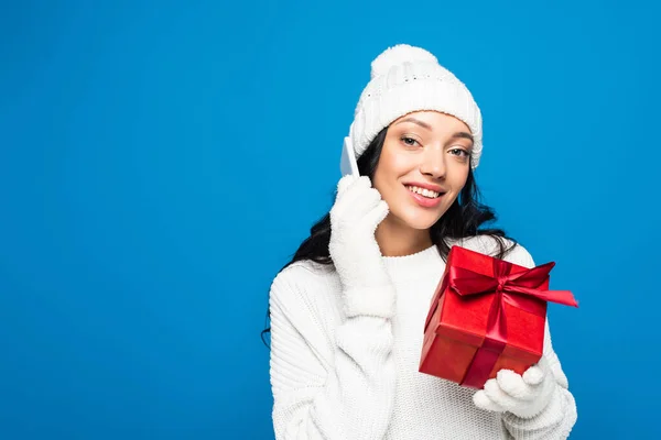 Happy woman in knitted hat and gloves holding gift box and talking on smartphone isolated on blue — Stock Photo