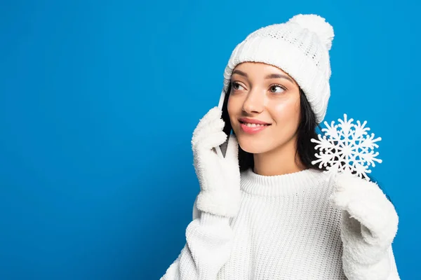 Felice donna in maglia cappello e guanti tenendo fiocco di neve decorativo e parlando su smartphone isolato su blu — Foto stock