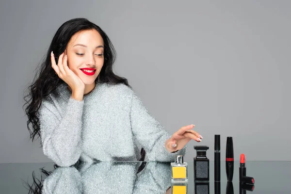 Mulher feliz com lábios vermelhos perto de garrafas com perfume e cosméticos decorativos isolados em cinza — Fotografia de Stock