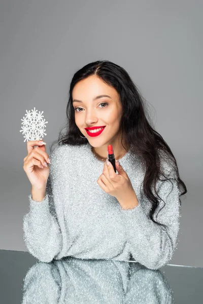 Pleased woman with red lips holding decorative snowflake and lipstick isolated on grey — Stock Photo