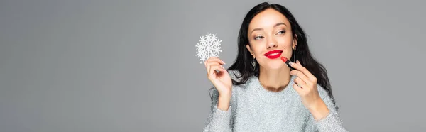 Pleased woman with red lips holding decorative snowflake and applying lipstick isolated on grey, banner — Stock Photo